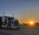 Semi-truck and dump trailer leaving a farm on a countryside road with sunrise in the background